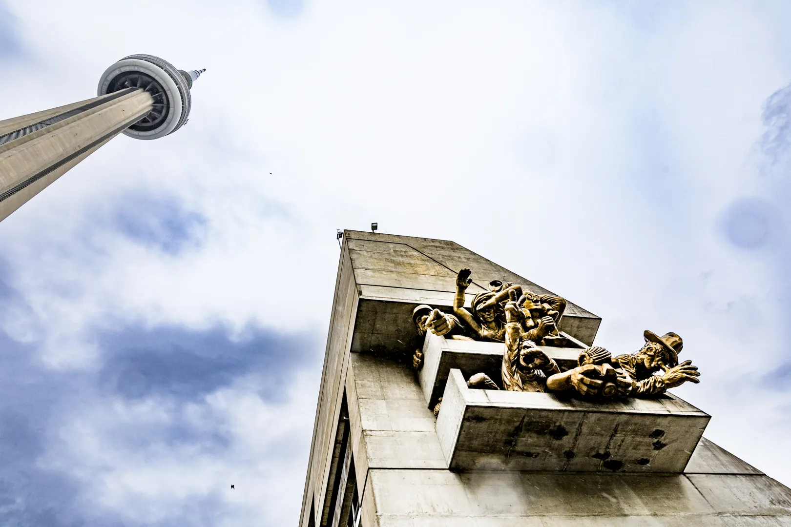 A photo of the CN Tower and a neighbouring building with human face statues coming out of it. Ein Foto des CN Towers und eines benachbarten Gebäudes mit menschlichen Gesichtsstatuten, die daraus herausragen.